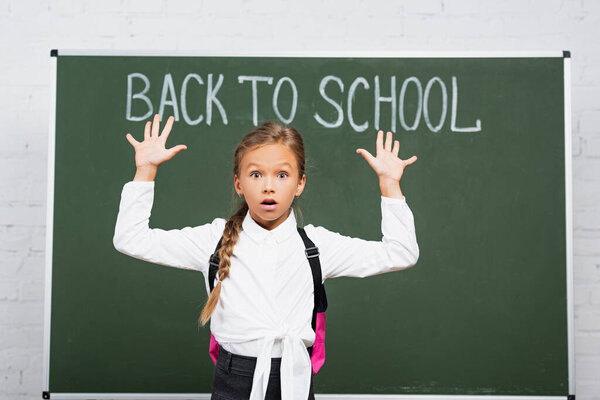 shocked schoolgirl standing with raised hands near chalkboard with back to school inscription