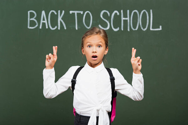 shocked schoolgirl holding crossed fingers near back to school inscription on chalkboard