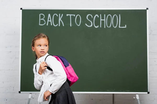 skeptical schoolgirl with backpack looking back near chalkboard with back to school inscription