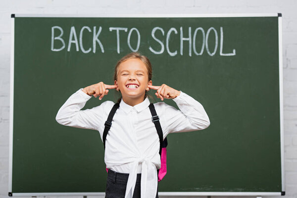 displeased schoolgirl plugging ears with fingers near chalkboard with back to school inscription