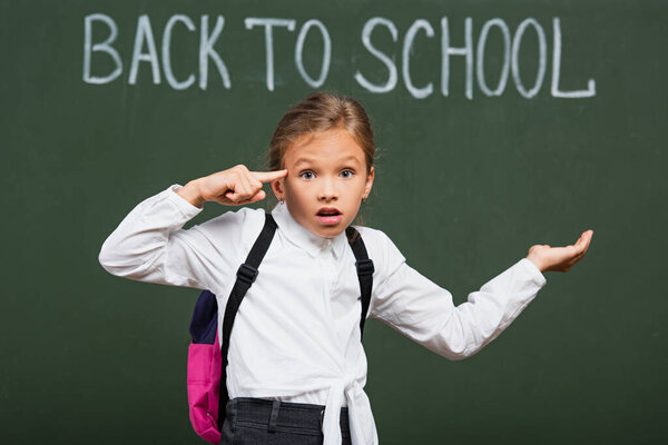 selective focus of discouraged schoolgirl showing crazy gesture while standing with open arm near back to school inscription on chalkboard