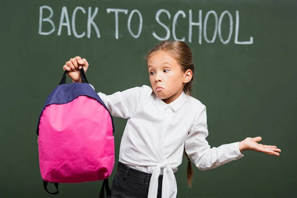 Confusa Colegiala Mostrando Gesto Encogimiento Mientras Sostiene Mochila Cerca Pizarra — Foto de Stock