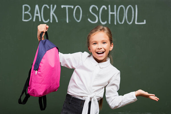 joyful schoolgirl with open arm holding backpack near chalkboard with back to school inscription