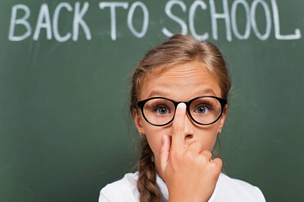 selective focus of surprised schoolgirl touching eyeglasses while looking at camera near back to school lettering on chalkboard