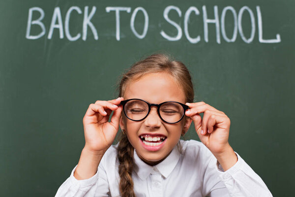 selective focus of excited schoolgirl laughing with closed eyes and touching eyeglasses near chalkboard with back to school inscription
