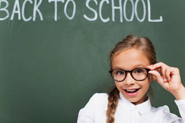 selective focus of cheerful schoolgirl touching eyeglasses near chalkboard with back to school lettering