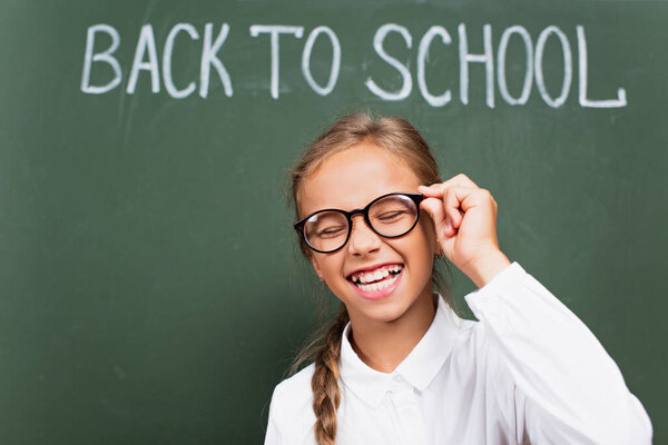 excited schoolgirl laughing with closed eyes and touching eyeglasses near chalkboard with back to school lettering