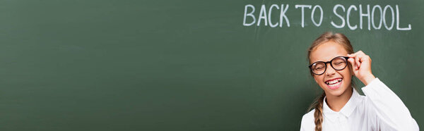 panoramic crop of excited schoolgirl laughing with closed eyes and touching eyeglasses near chalkboard with back to school lettering