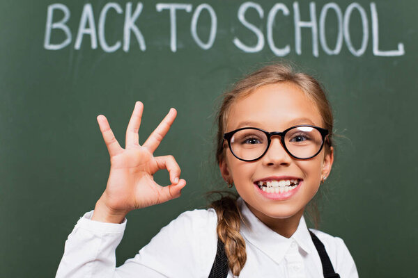 selective focus of excited schoolgirl in eyeglasses showing thumb up near chalkboard with back to school text