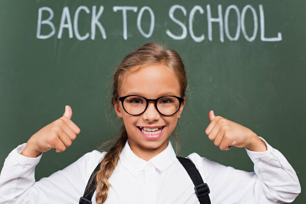 selective focus of happy schoolgirl in eyeglasses showing thumbs up near chalkboard with back to school lettering