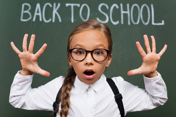 selective focus of schoolgirl in eyeglasses showing scaring gesture near chalkboard with back to school lettering