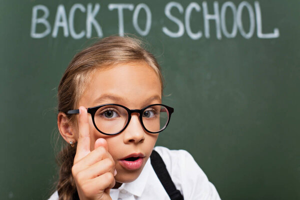 selective focus of adorable schoolgirl in eyeglasses showing idea gesture near chalkboard with back to school text