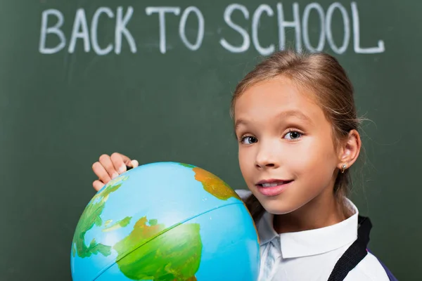 笑顔の女子高生が世界を握り黒板に学校の文字が書かれたカメラを見て — ストック写真
