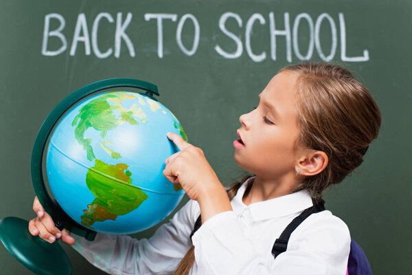 selective focus of attentive schoolgirl pointing with finger at globe near back to school lettering on chalkboard