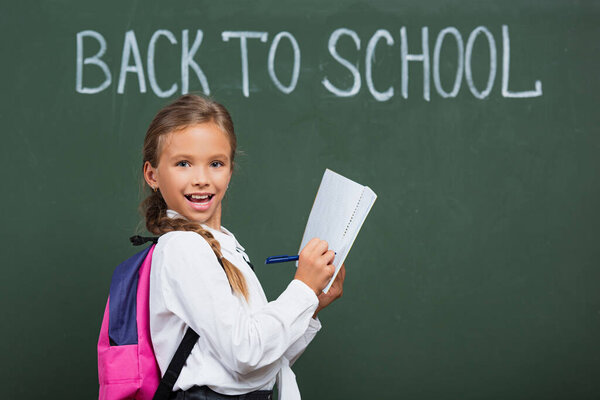 happy schoolgirl with backpack writing in notebook near chalkboard with back to school lettering