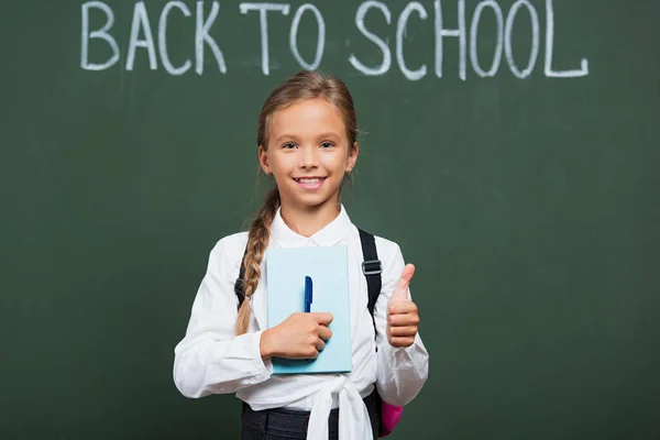 一个快乐的女学生 手里拿着书和笔 在黑板旁边竖起大拇指 背对着学校的字母 — 图库照片