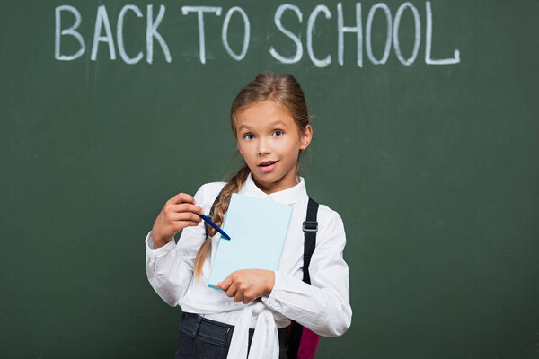 surprised schoolgirl pointing with pen at book near chalkboard with back to school inscription