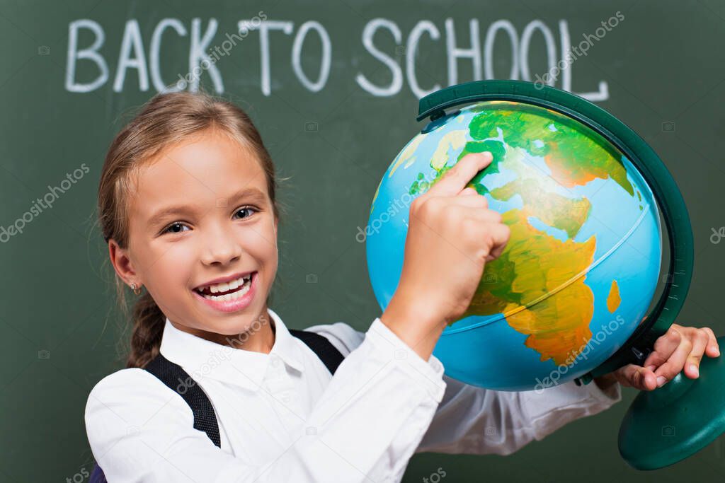 selective focus of happy schoolgirl pointing with finger at globe near back to school inscription on chalkboard