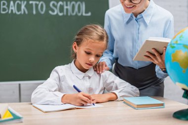 Gülümseyen öğretmenin, masada oturan ve deftere yazan öğrenciye bakışının seçici odak noktası. 