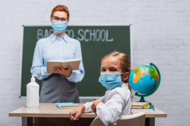 Koruyucu maskeli bir kızın kameraya baktığı seçici odak noktası ve kitap ile yazı tahtasının yanındaki öğretmen.