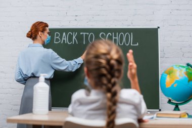 Yükselmiş elli bir kız öğrencinin arka görünüşü ve okula geri dönen karatahtaya yazı yazan tıp maskeli bir öğretmen.