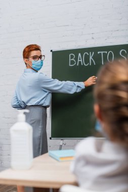 Okul kızlarının arka planı ve tıbbi maskeli öğretmenin tahtaya yazı yazması. 