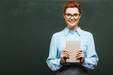 Gözlüklü mutlu öğretmen karatahtanın yanında dururken elinde kitap tutuyor.  