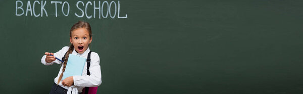 horizontal image of excited schoolgirl pointing with pen at book near chalkboard with back to school inscription  