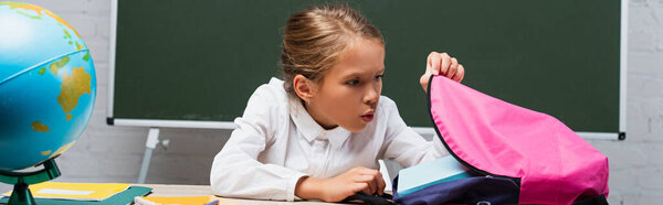 panoramic shot of surprised schoolgirl looking into backpack while sitting at desk near globe and chalkboard 