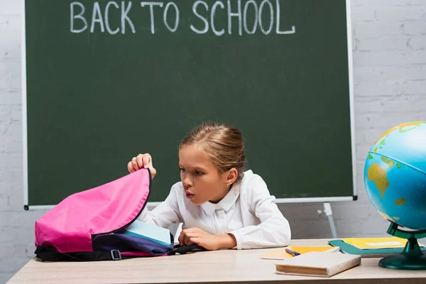 Colegiala Sorprendida Mirando Mochila Mientras Está Sentado Escritorio Cerca Globo — Foto de Stock