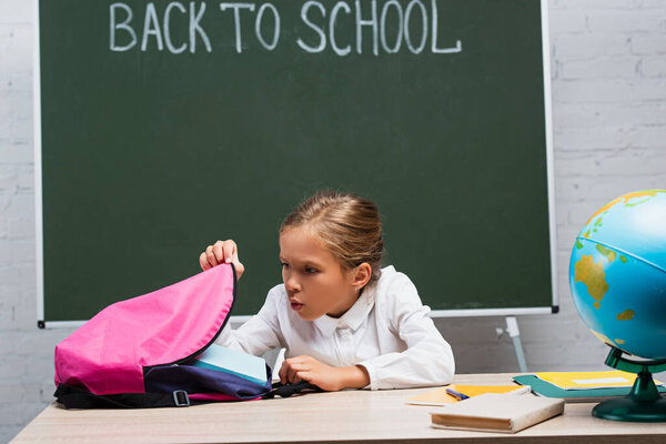 surprised schoolgirl looking into backpack while sitting at desk near globe and chalkboard with back to school lettering