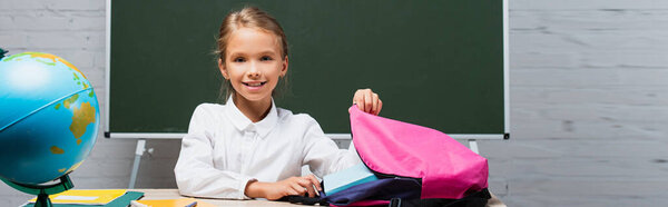 smiling schoolgirl taking books from backpack while sitting at desk near globe and chalkboard