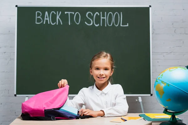 Colegiala Sonriente Tomando Libros Mochila Mientras Está Sentado Escritorio Cerca —  Fotos de Stock