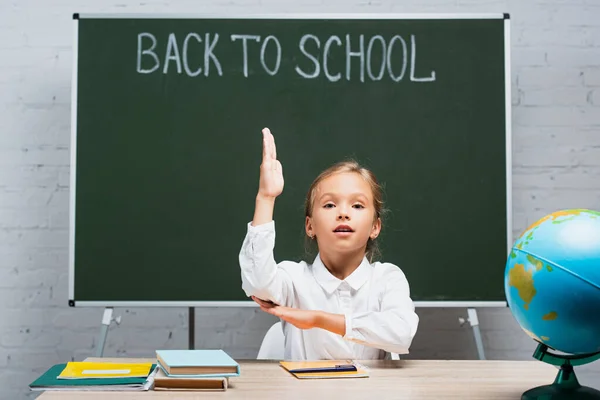 Adorable Schoolgirl Raising Hand While Sitting Desk Globe Chalkboard Back — Stock Photo, Image