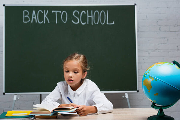 attentive schoolgirl reading book while sitting at desk near globe and chalkboard with back to school inscription   