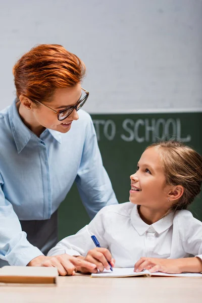 Happy Schoolgirl Looking Attractive Teacher While Writing Copy Book — Stock Photo, Image