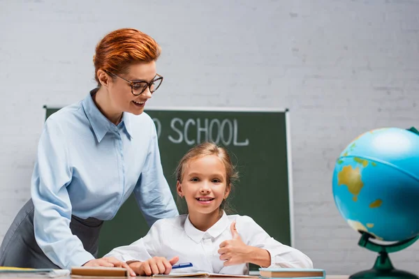 Leende Lärare Står Nära Glad Skolflicka Sitter Vid Skrivbordet Och — Stockfoto