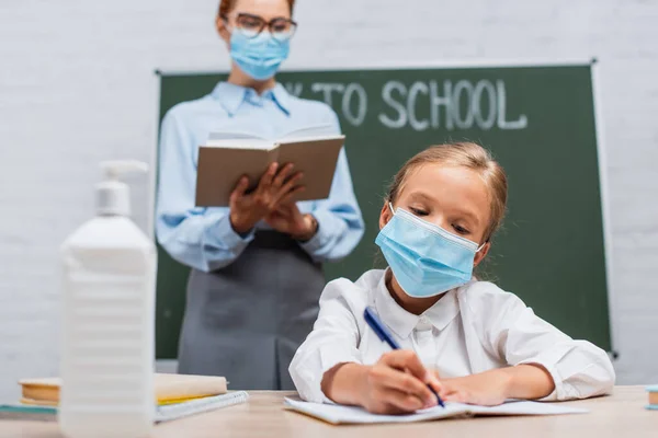 selective focus of schoolgirl in medical mask writing in notebook, and teacher standing near chalkboard with book