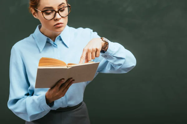 Attentive Teacher Eyeglasses Pointing Finger Open Book While Standing Chalkboard — Stock Photo, Image