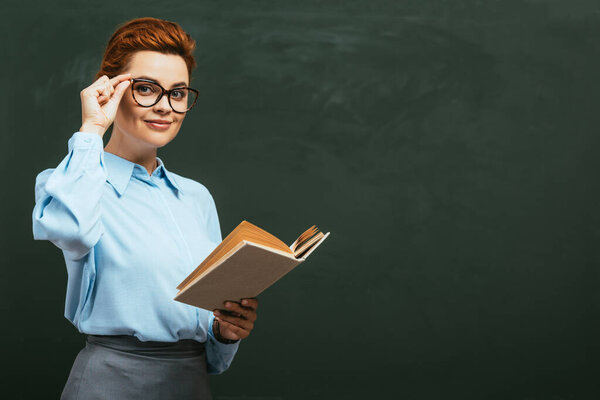 улыбающийся учитель трогает очки, стоя рядом с доской с открытой книгой