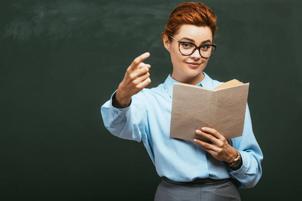 улыбающийся учитель держит открытую книгу и показывает прийти сюда жест, стоя рядом с доской 