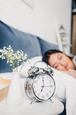 Alarm saatinin seçici odağı ve vazoda çiçekler komodinin üzerinde ve Asyalı bir kadın sabah yatağında uyuyor. 