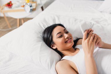 Evde yatakta akıllı telefon kullanan pozitif Asyalı kadının yüksek açılı görüntüsü 