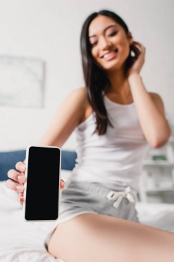 Gülümseyen Asyalı kadının seçici odak noktası yatakta boş ekranla akıllı telefon göstermesi. 