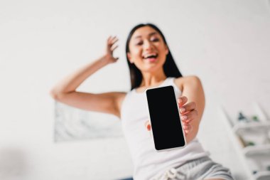 Evde boş ekranlı akıllı telefon gösteren neşeli Asyalı kadının seçici odak noktası 