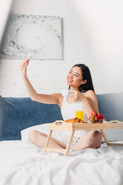 Gülümseyen Asyalı kadının selfie çektiği ve kahvaltının yanında kahvaltını tuttuğu seçmeli odak noktası. 