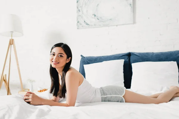 魅力的なアジアの女の子笑顔でカメラに横たわっている間にベッド — ストック写真