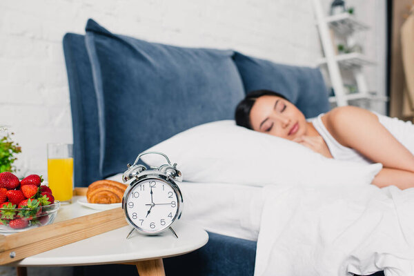 Selective focus of alarm clock and breakfast on bedside table and asian woman sleeping on bed 