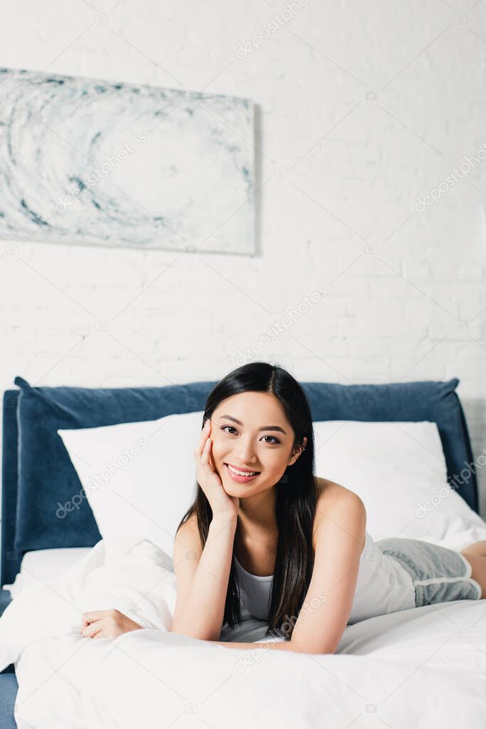 Young asian woman with hand near head smiling at camera on bed 