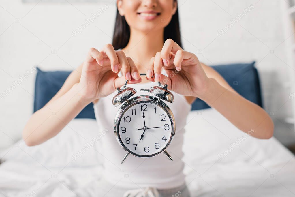 Selective focus of smiling woman holding alarm clock in bedroom at morning 